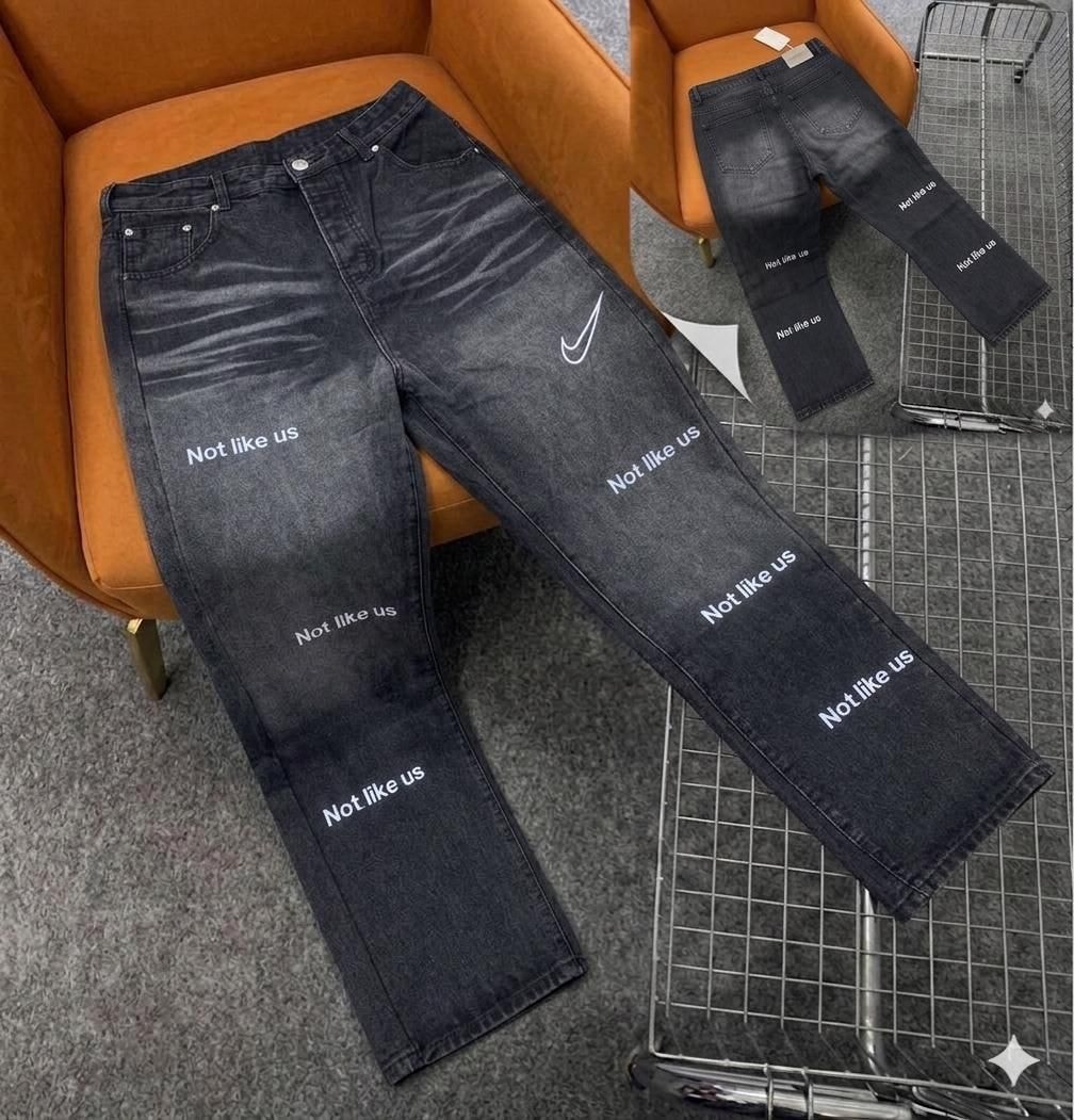 Dark wash baggy jeans with repeated 'Not like us' white text prints and swoosh logo on thigh.