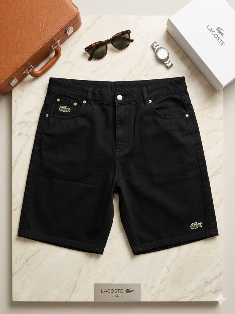 Black Lacoste denim jorts with green crocodile logos on marble surface with accessories.