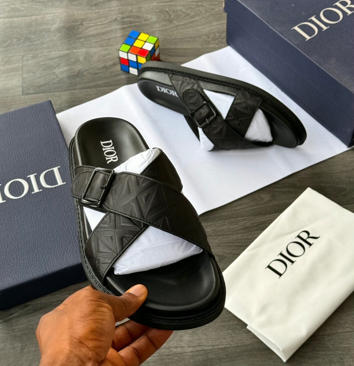 Hand holding black Dior cross strap slide with buckle and embossed pattern.