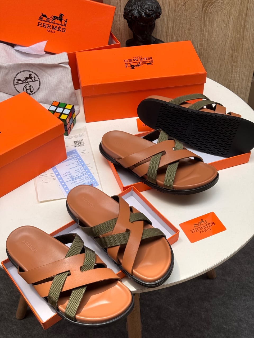 Brown and green strappy Hermes slides shown with boxes, Rubik's cube and accessories.