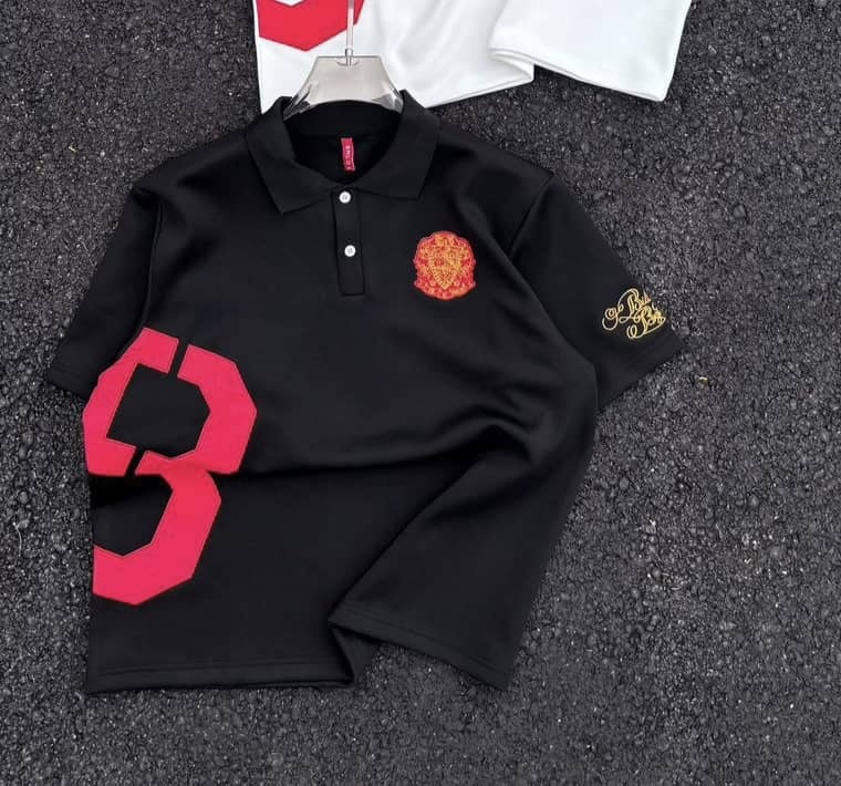 Black polo shirt with red number graphic, red-yellow chest emblem and gold sleeve embroidery, with white version in background.