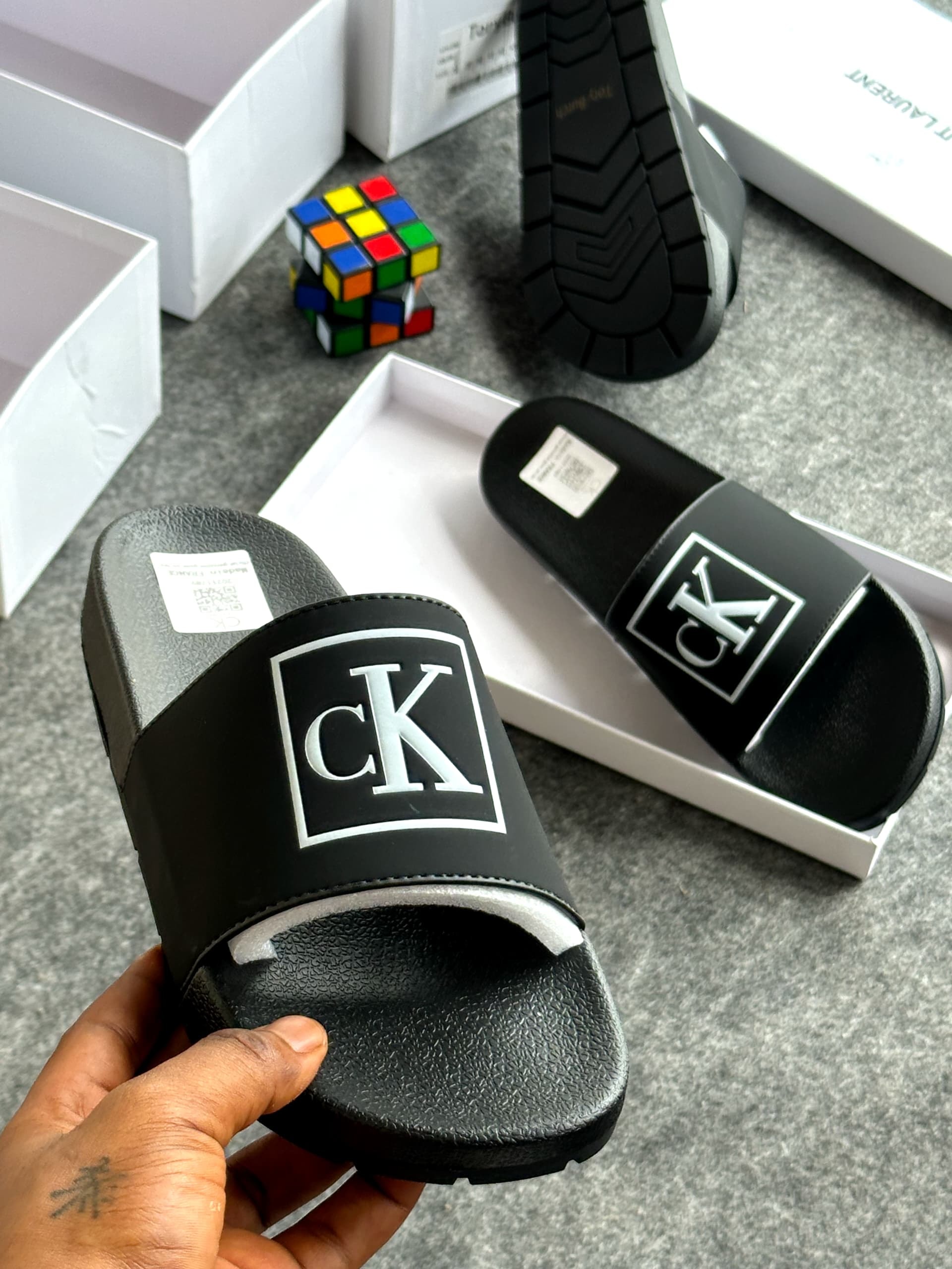 Black slide with CK logo in silver square, held displaying the design and background items.
