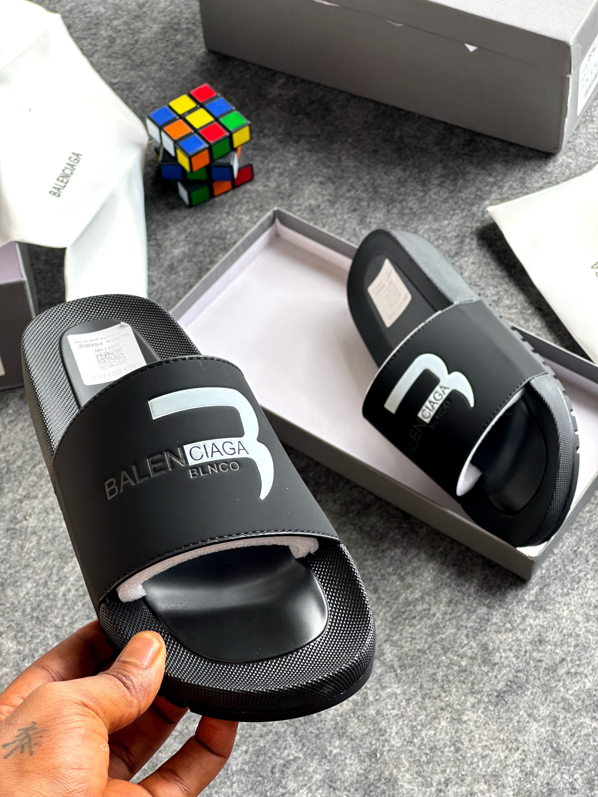 Black Balenciaga slide with large white logo held in hand displaying details and packaging.
