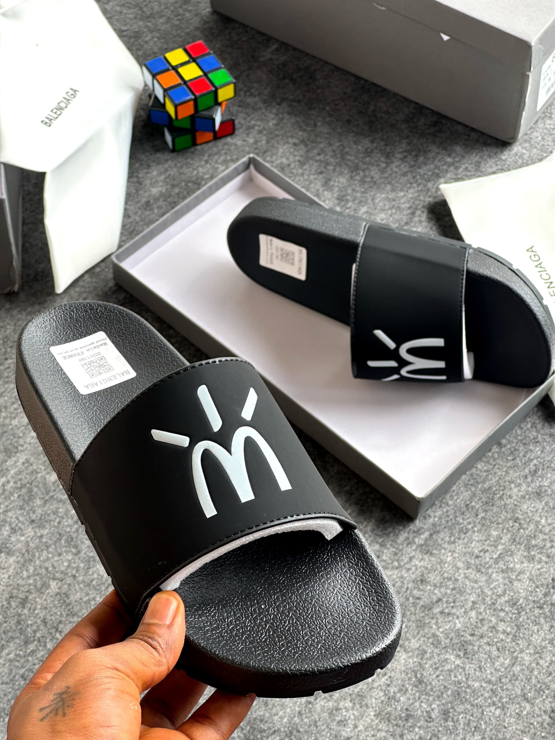 Black slide with white stylized logo on strap held in hand, with Balenciaga packaging and Rubik's cube visible.