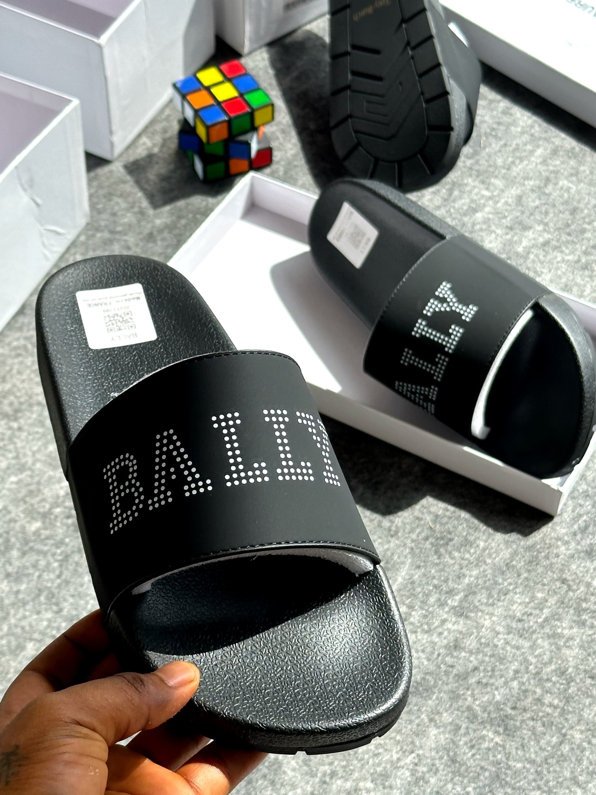 Black slide with BALLI logo in white dots on the strap, held in hand showing textured sole and packaging.