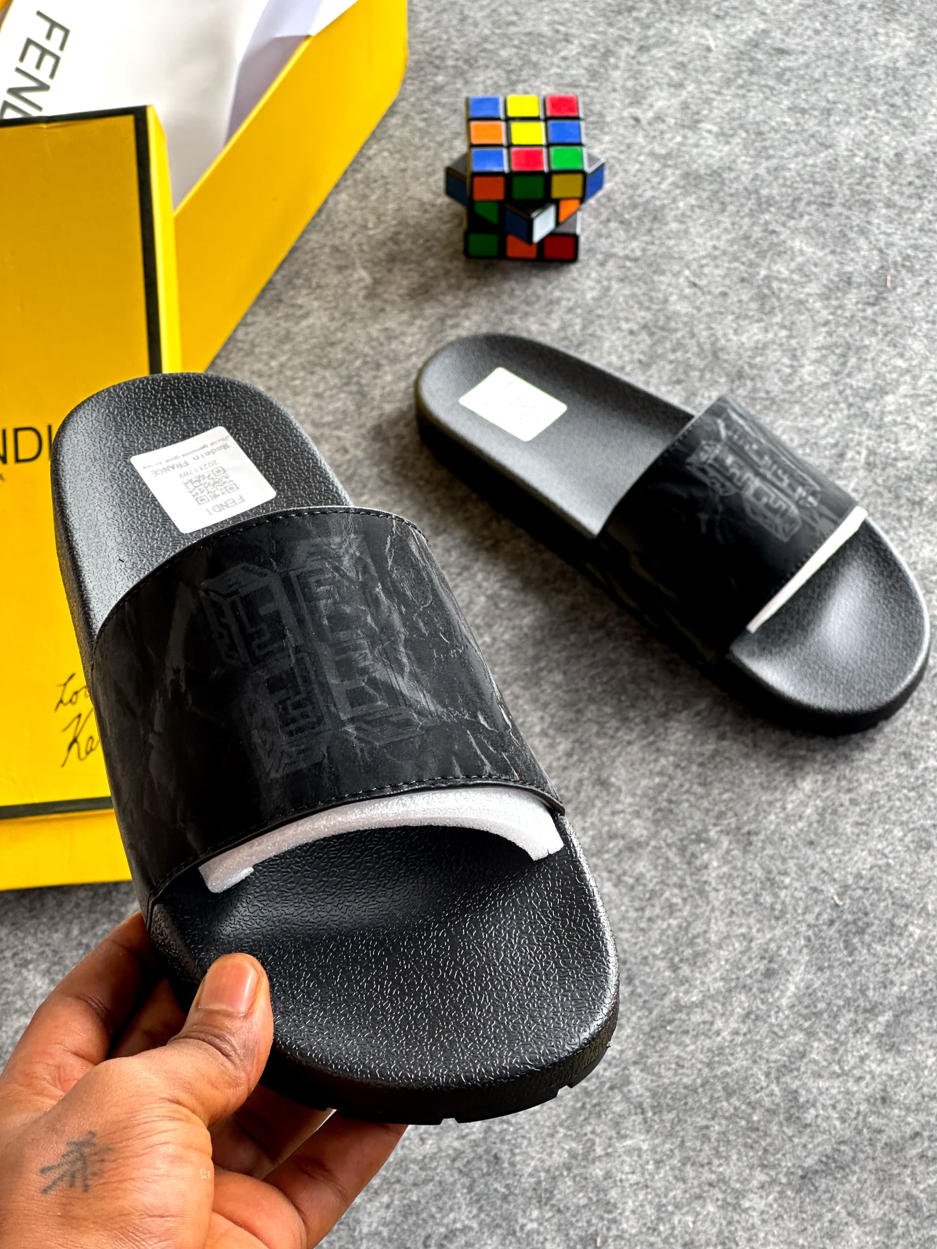 Black Fendi slides with embossed logo pattern held in front of yellow branded box.