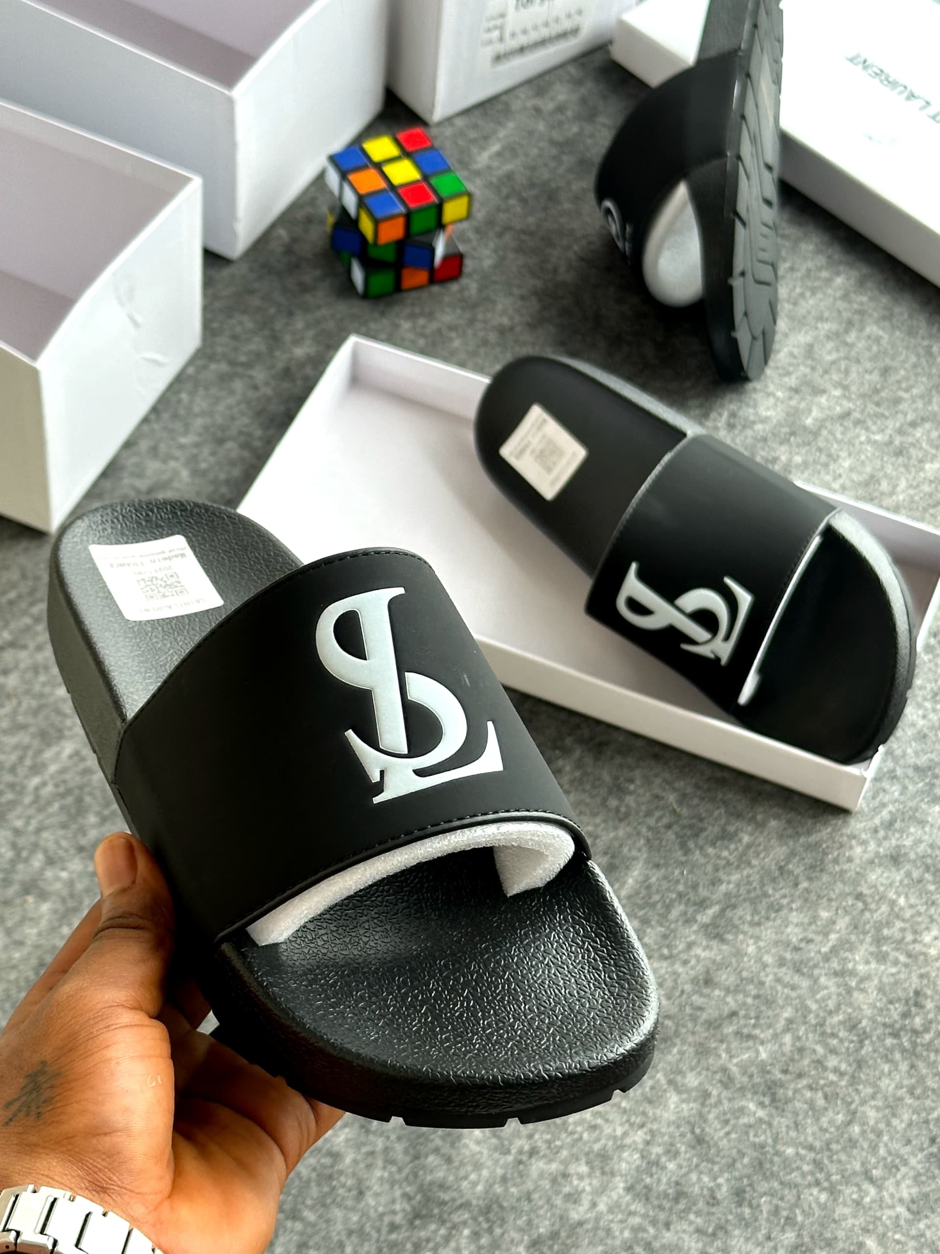 Black YSL slides with large silver logo shown with open box and accessories.
