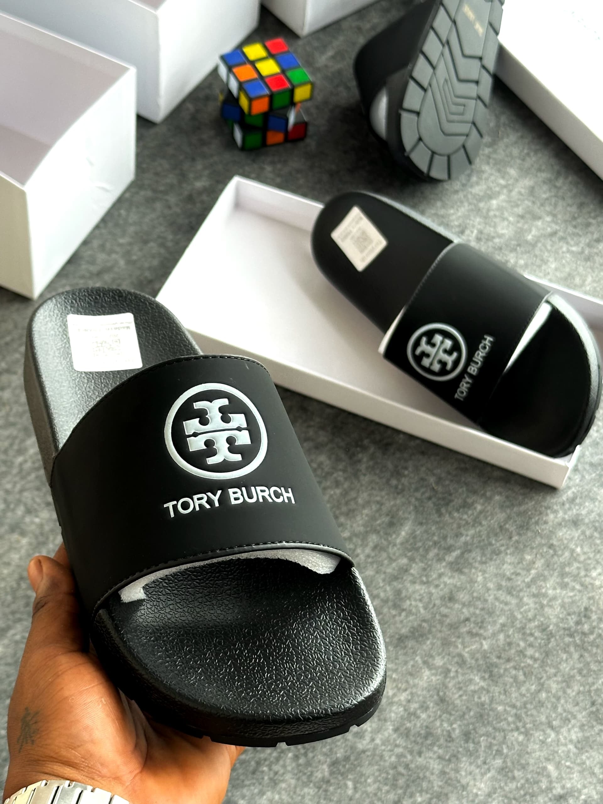 Black Tory Burch slides with prominent logo held above white product boxes.