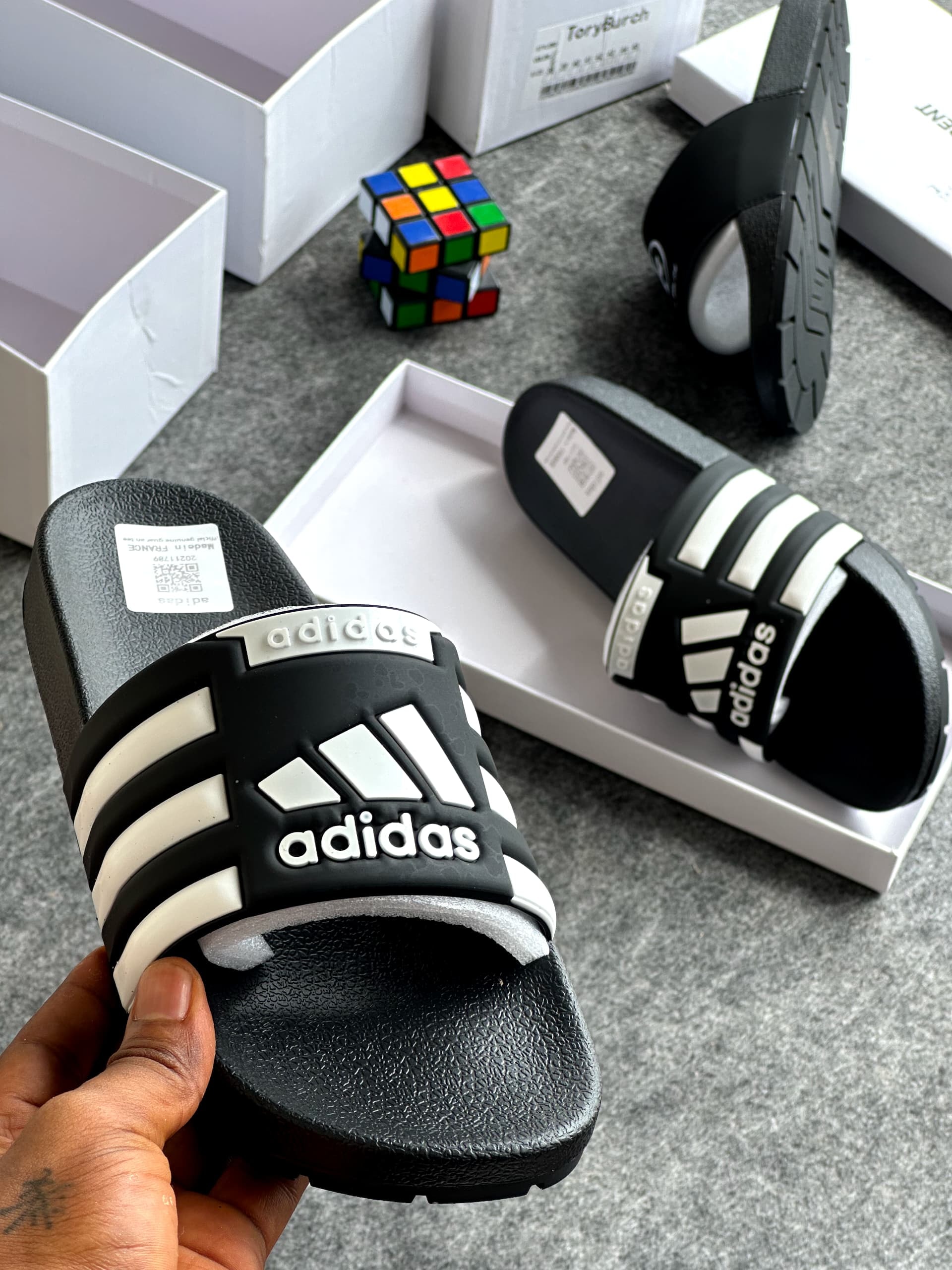 Black Adidas slides with white stripes and logo shown with product boxes and Rubik's cube.