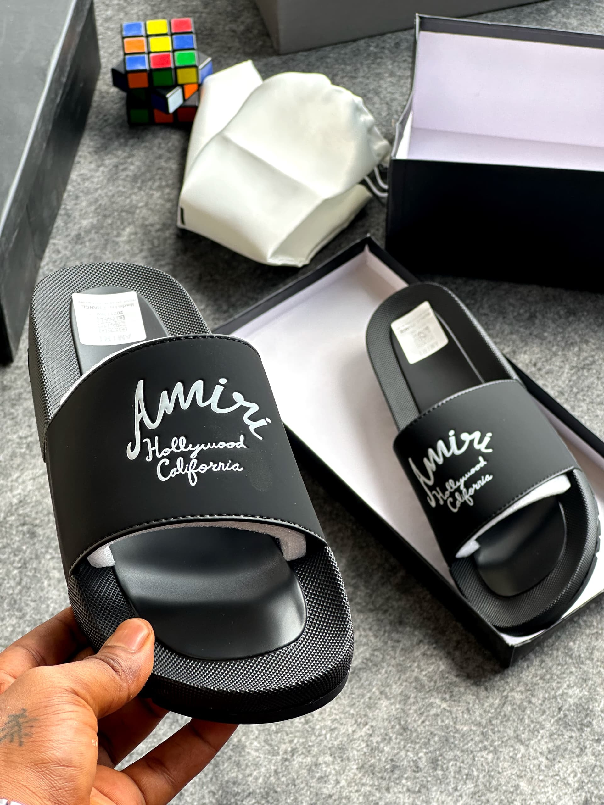 Black Amiri slides with white script logo held above open branded box and accessories.