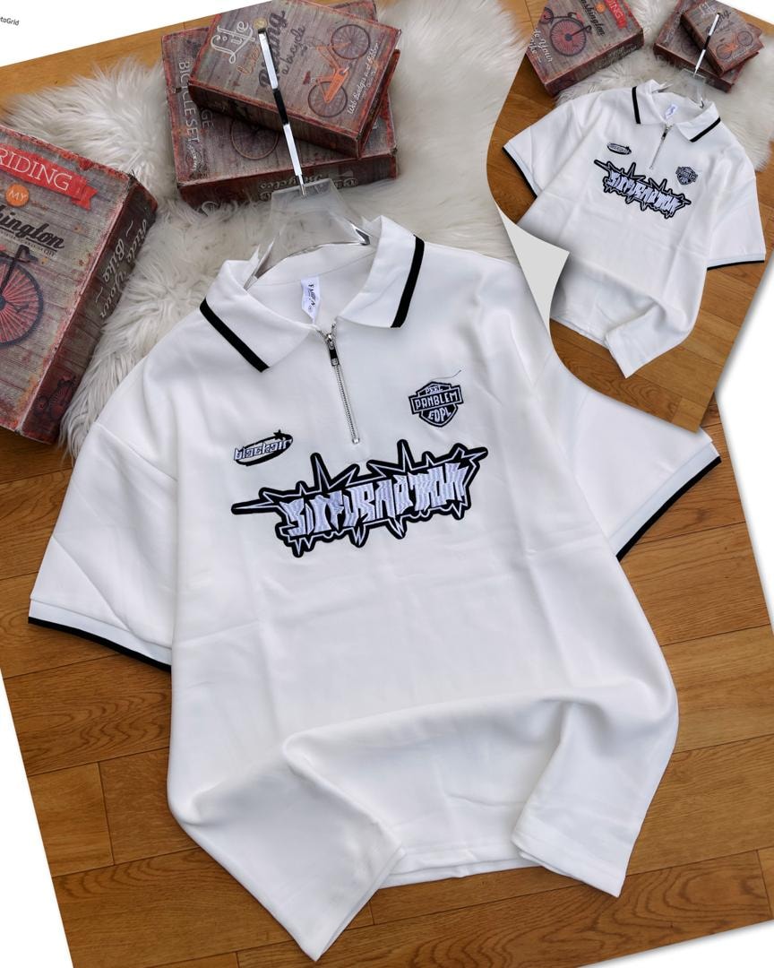 White zip collar polo shirt with graphic embroidery and patches on chest, shown on wooden surface with smaller inset view.