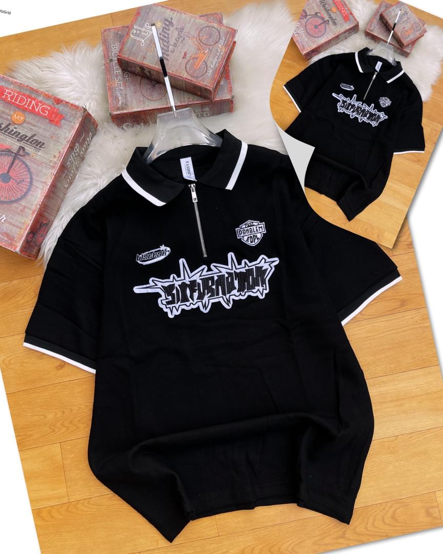 Black zip collar polo shirt with graphic embroidery and patches on chest, shown on wooden surface with smaller inset view.