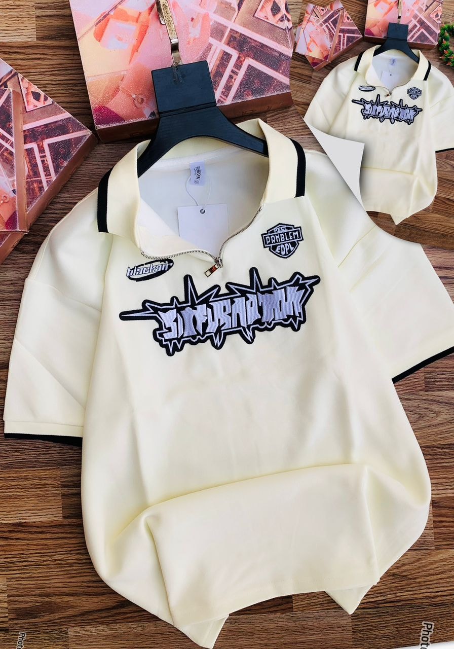 Cream zip collar polo shirt with graphic embroidery and patches on chest, shown on wooden surface with smaller inset view.