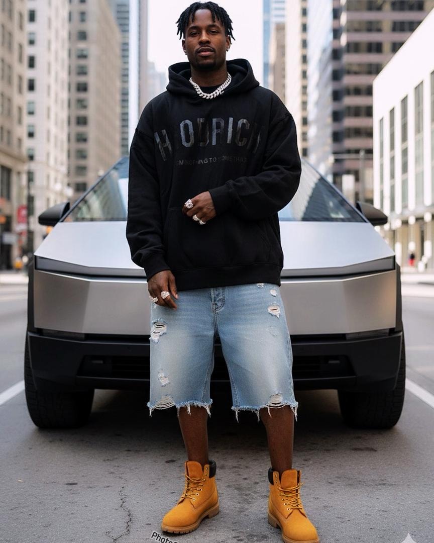 Man in black Hoodrich graphic hoodie, ripped denim shorts and yellow boots posing in front of silver vehicle.