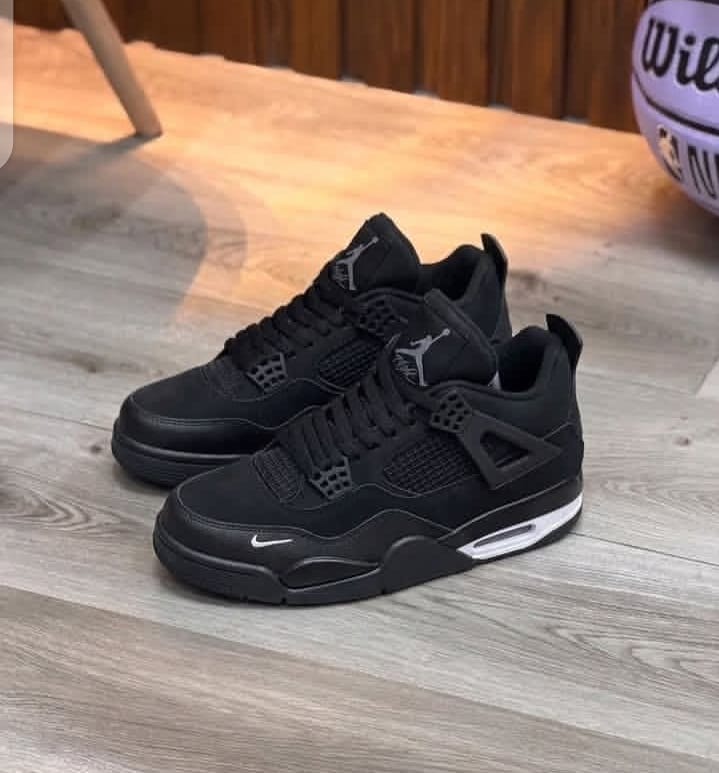 Pair of Nike Air Jordan 4 Black Cat sneakers placed on wooden floor with basketball in background.