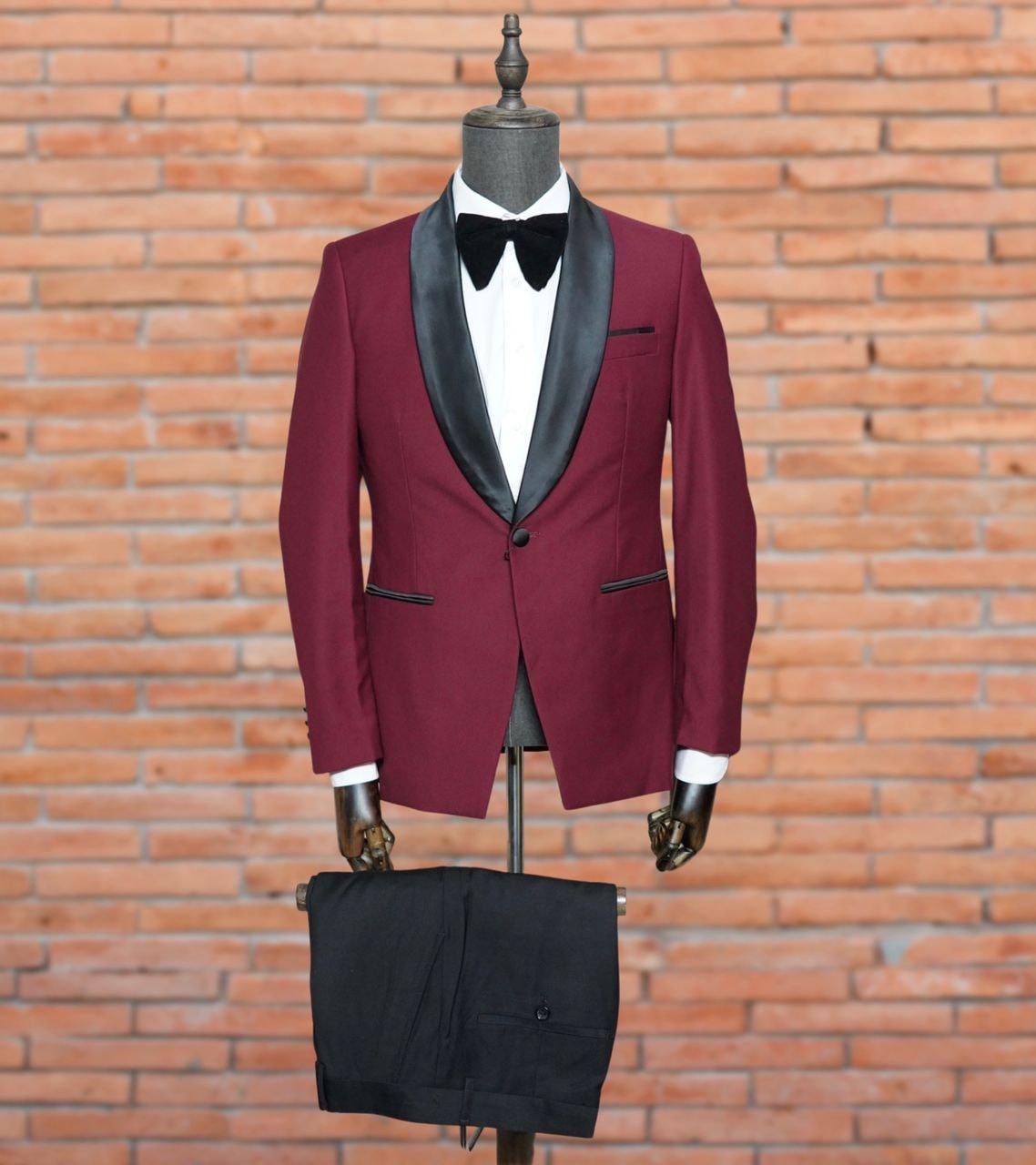 Burgundy tuxedo with black shawl lapels, bow tie, white shirt and black trousers on mannequin against brick wall.