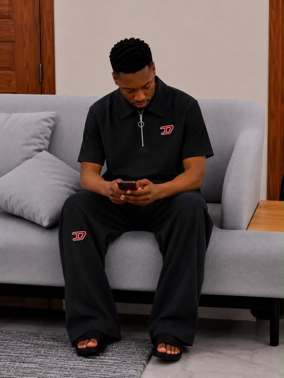 Man seated on gray sofa wearing black ribbed two-piece set with red D logo on chest and leg.