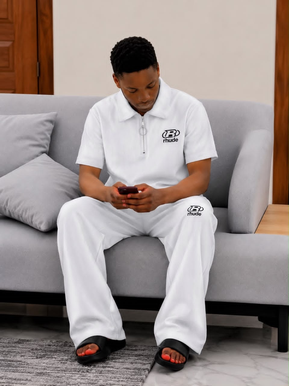 Man seated on gray sofa wearing white ribbed two-piece set with black Rhude logo on chest and leg.