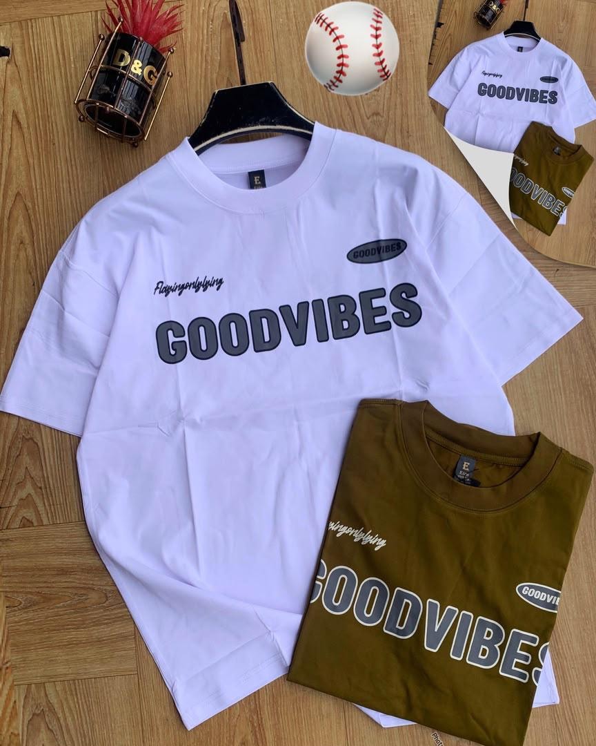 White and olive Good Vibes printed t-shirts on wooden surface with baseball and plant props.