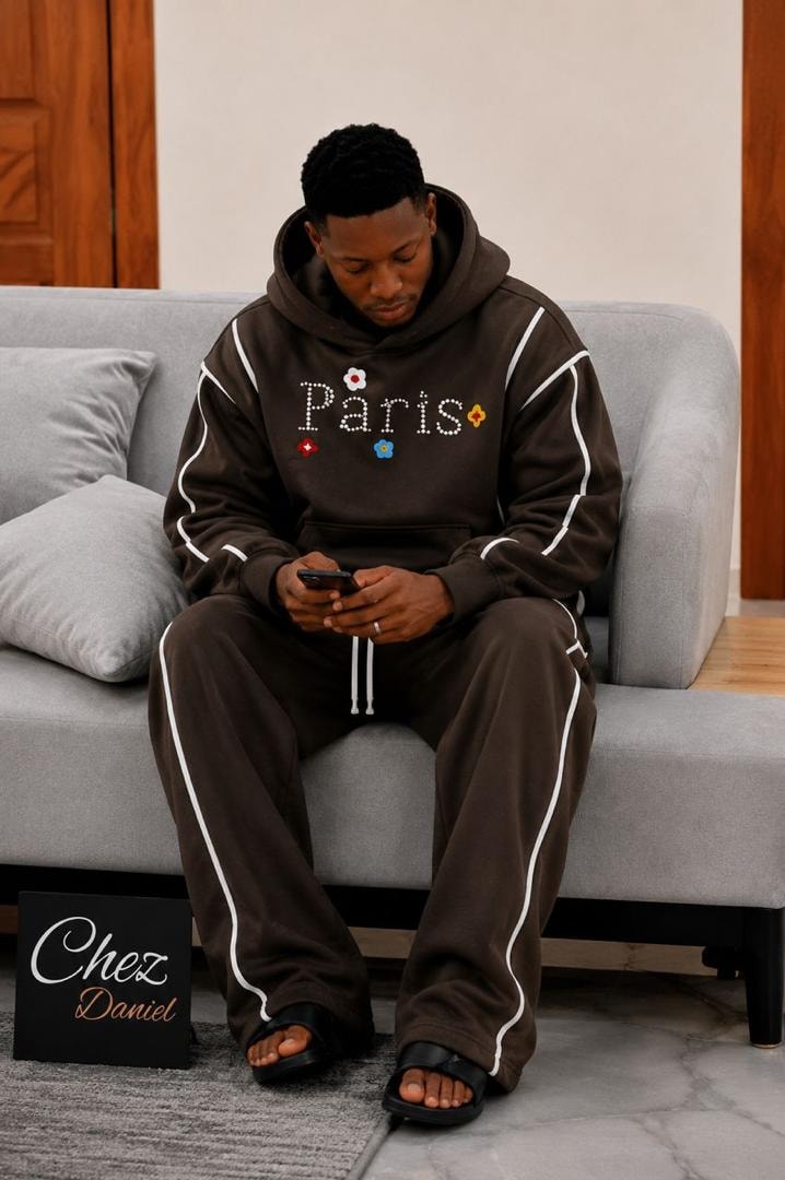 Model wearing brown Paris beaded two-piece hoodie and pants set with white piping.