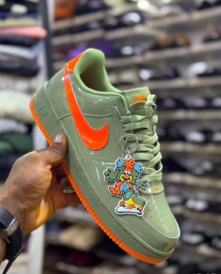 Hand holding green and orange Nike Air Force 1 sneaker with clown keychain in store setting.