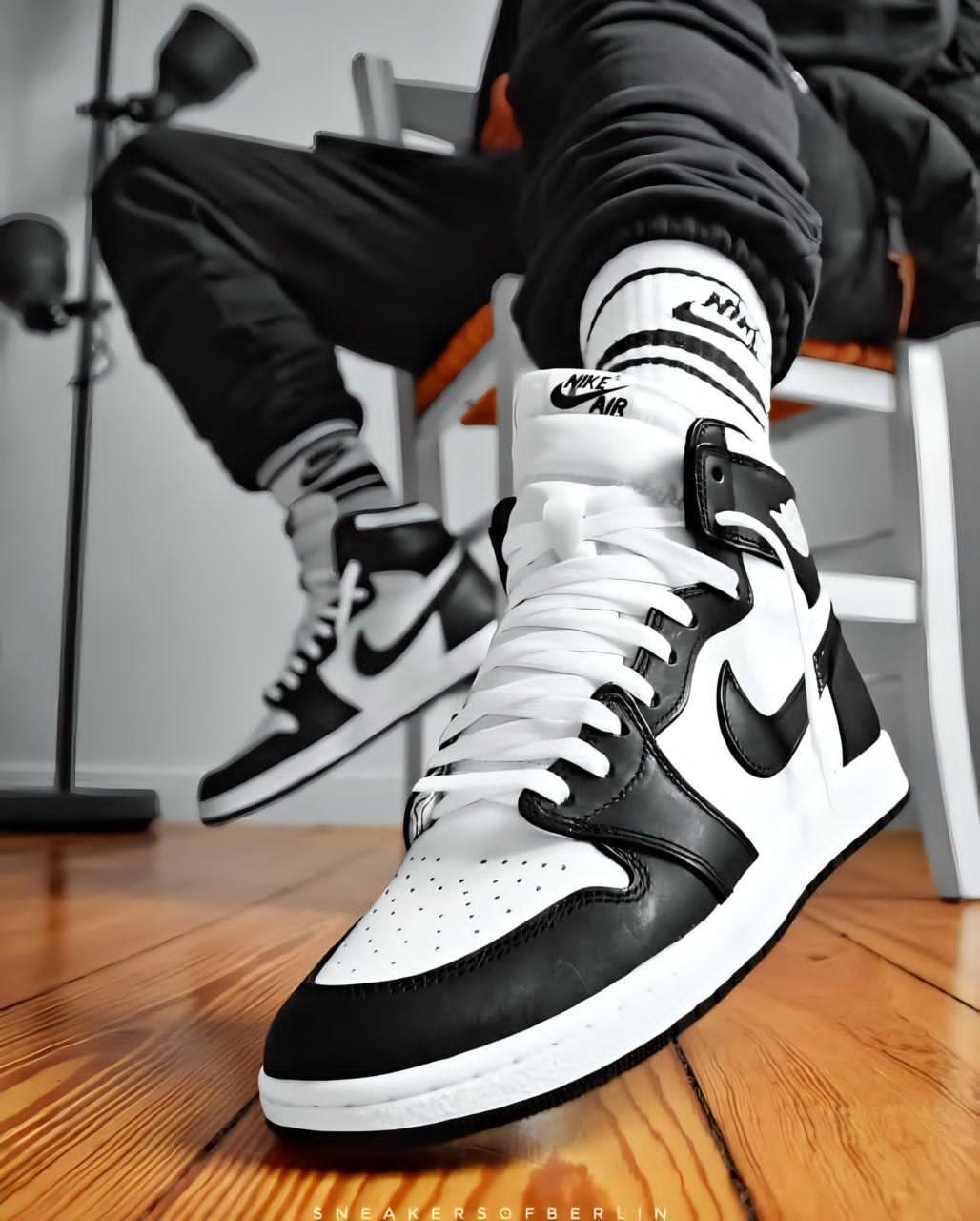 Black and white Air Jordan 1 High sneakers worn with black pants on wooden floor.