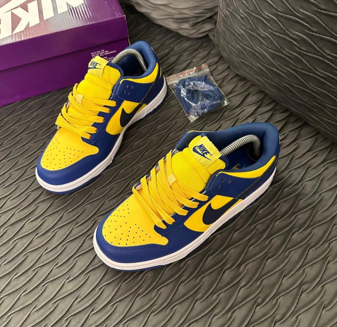 Blue and yellow Nike Dunk Low sneakers on gray textured surface with purple box.
