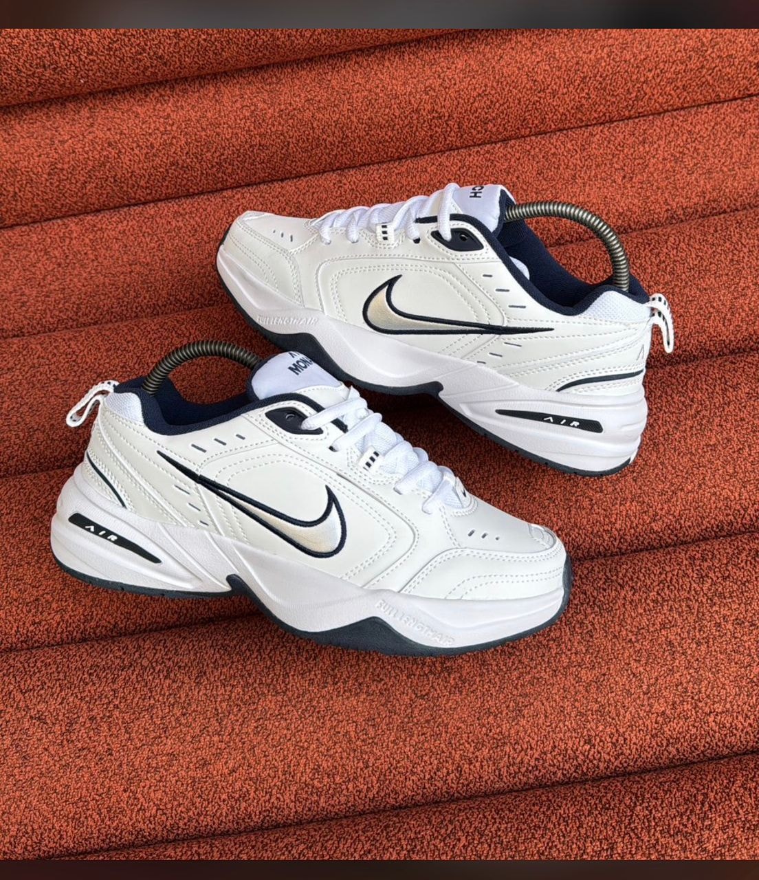 White Nike Air Monarch sneakers with black swoosh on orange textured surface.