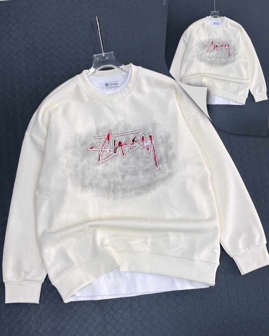 Cream Stussy sweatshirt with red script graphic on chest and back, shown on hanger.