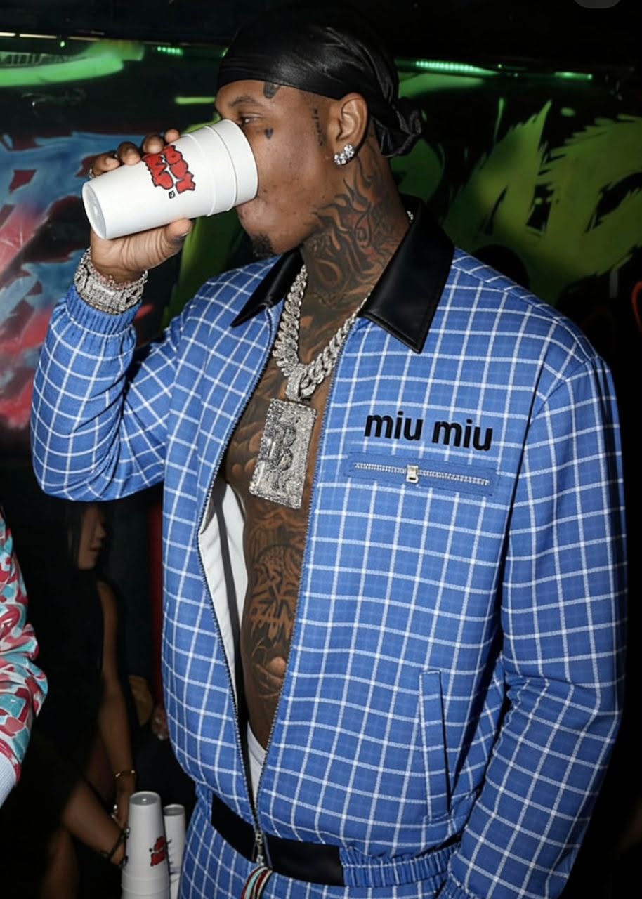 Man in blue grid miu miu jacket drinking from a cup, showing side profile.