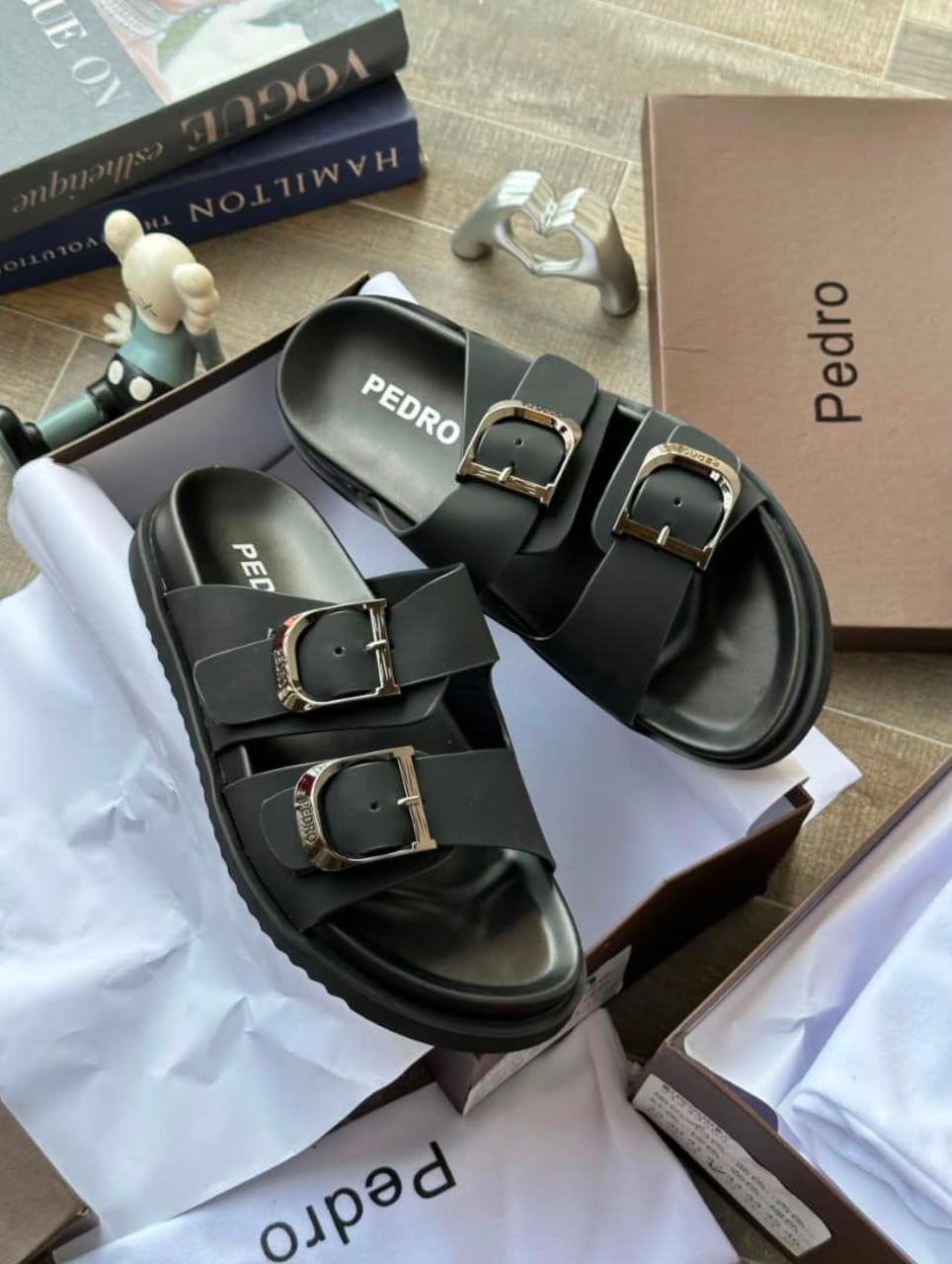 Black Pedro double-buckle slides in box with books and decorative items.