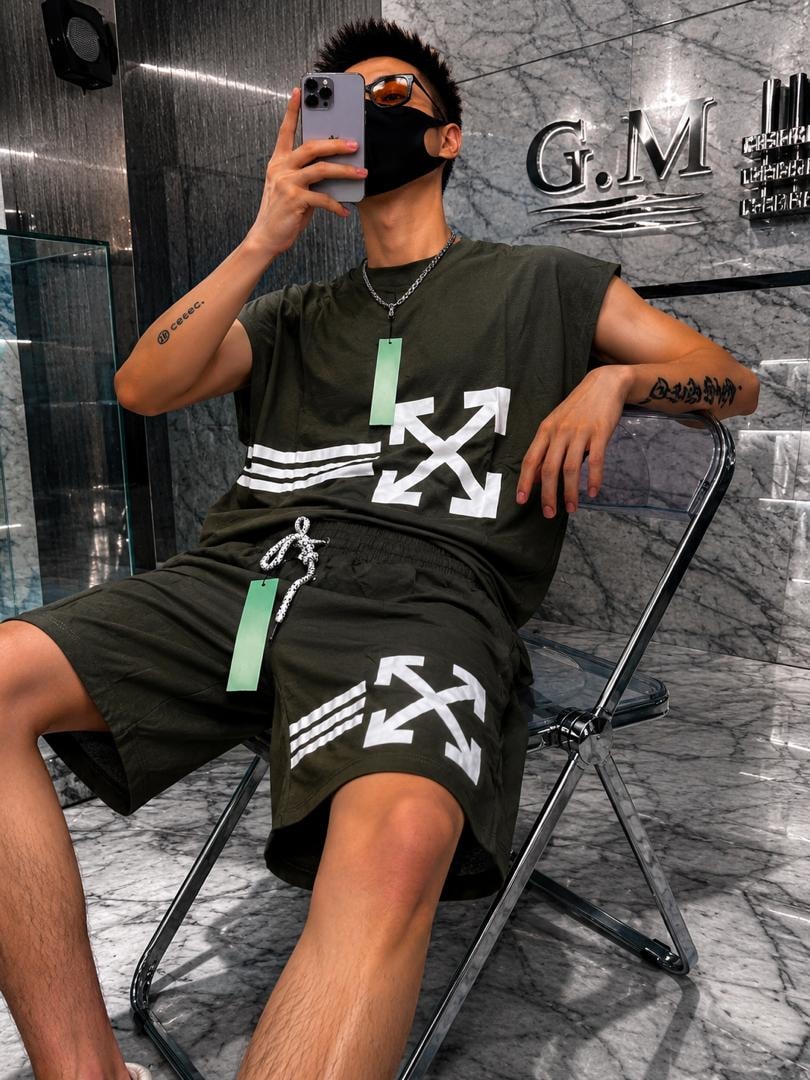 Man seated wearing olive green sleeveless top and matching shorts with white arrow graphics.