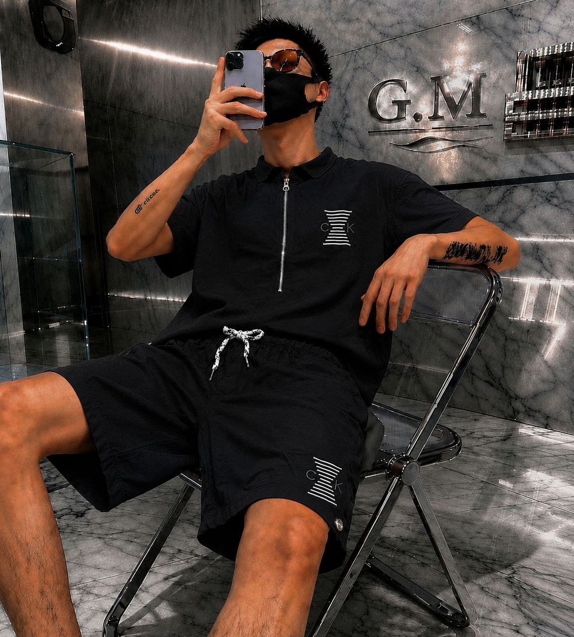 Man seated wearing black short-sleeve zip polo shirt and matching shorts with white COK logo.