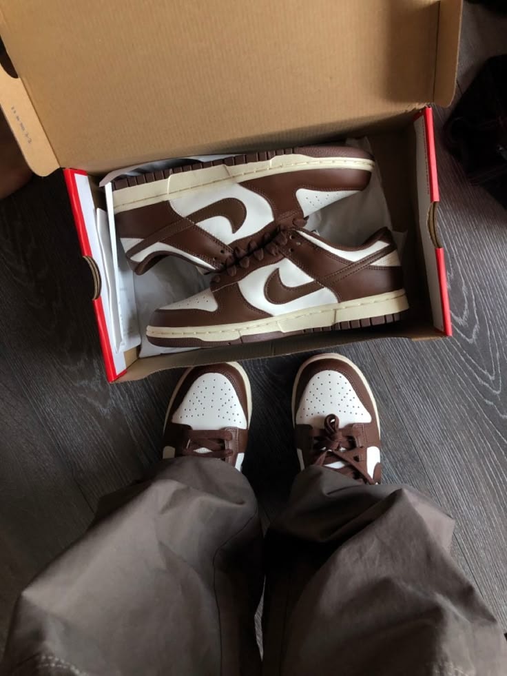 Nike Dunk Low brown and white sneakers inside open box with person wearing brown trousers.