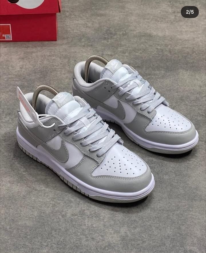 Pair of grey and white Nike Dunk Low sneakers on grey floor with red box in background.
