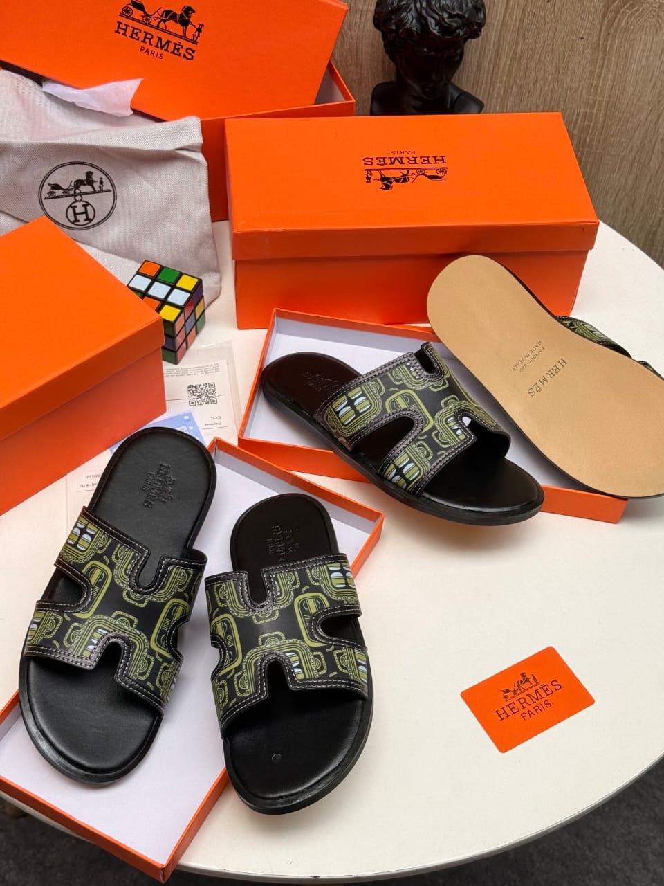 Black Hermes slides with green pattern on strap shown with orange boxes and dust bag.