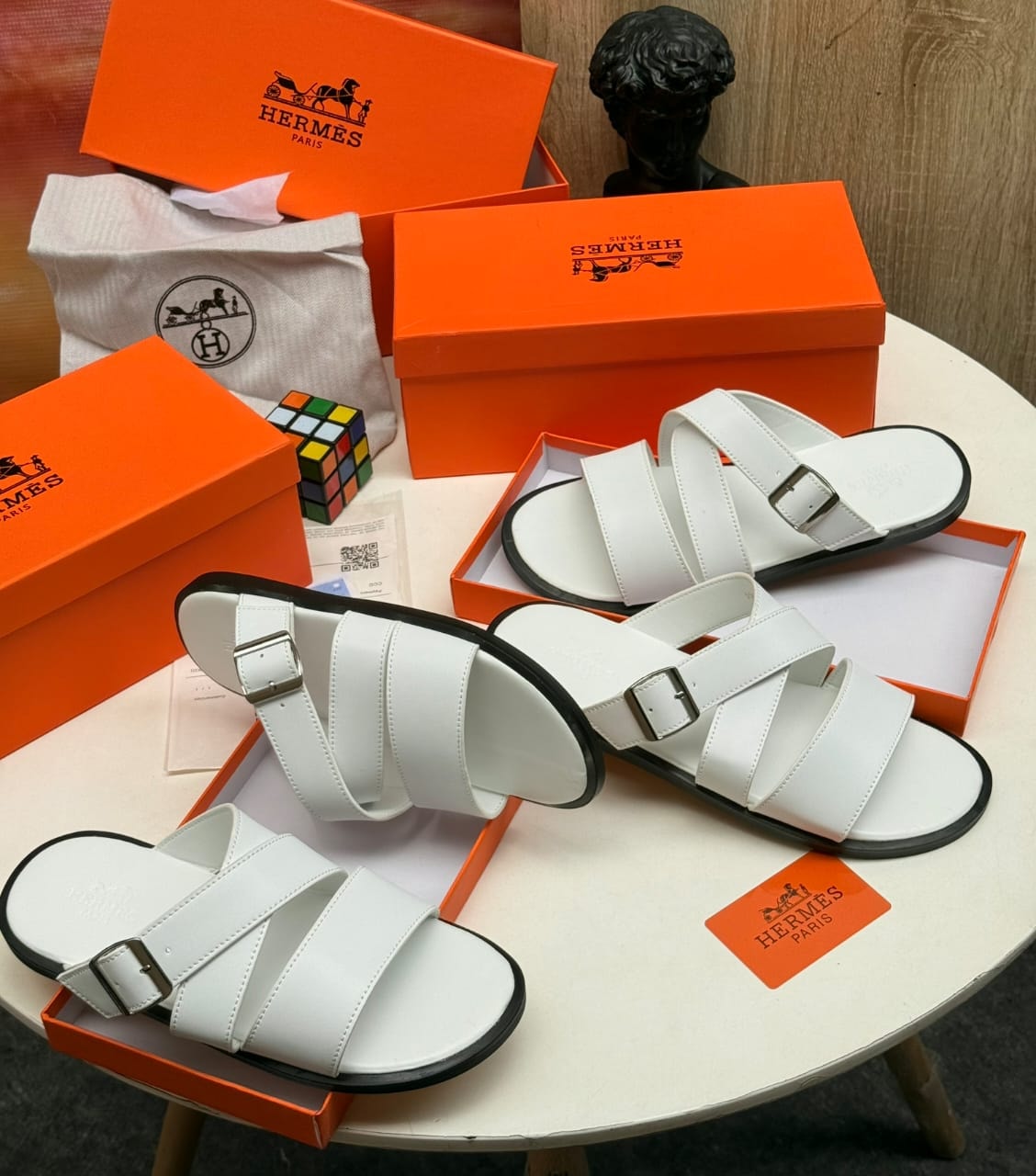White Hermes multi-strap slides with buckles displayed with orange packaging.