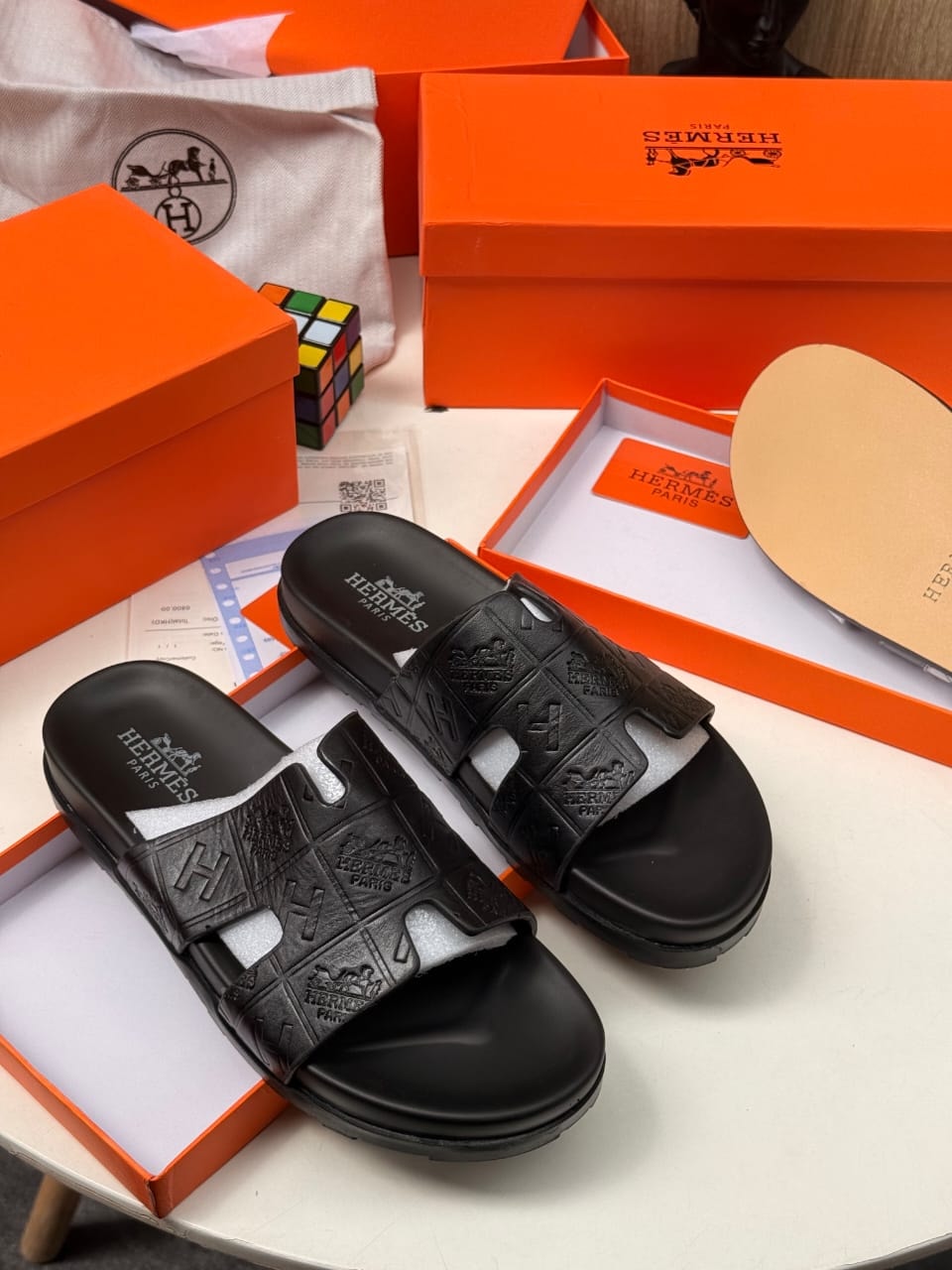 Black Hermes slides with embossed H and logo pattern in orange box setup.