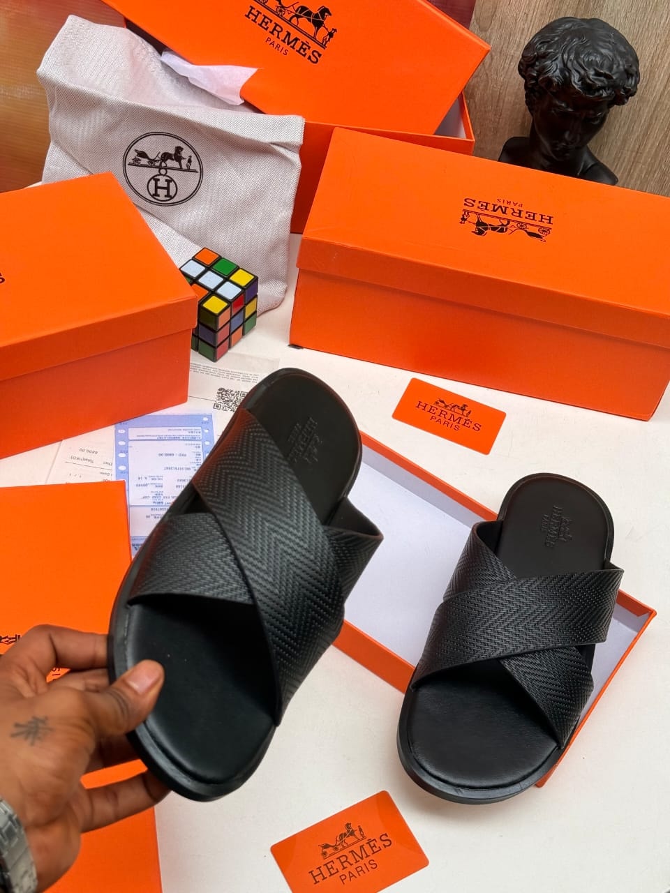 Black Hermes cross-strap slides with textured pattern and branding shown with boxes.