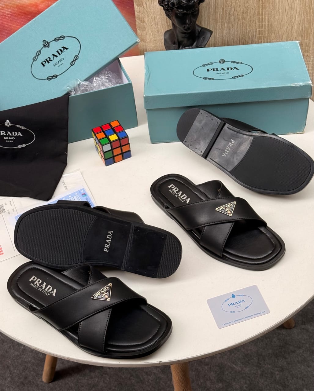 Black Prada double-strap slides with gold triangle logo displayed with branded box and accessories.