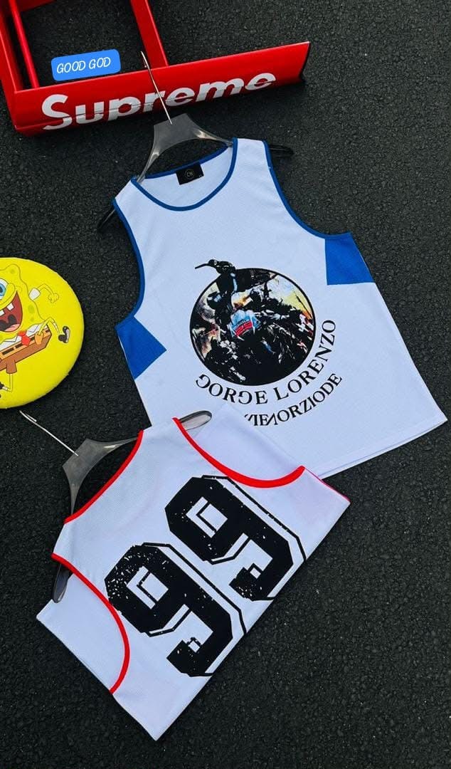 Flat lay of white armless tank with large 99 print and red trim alongside graphic tank on dark surface with props.