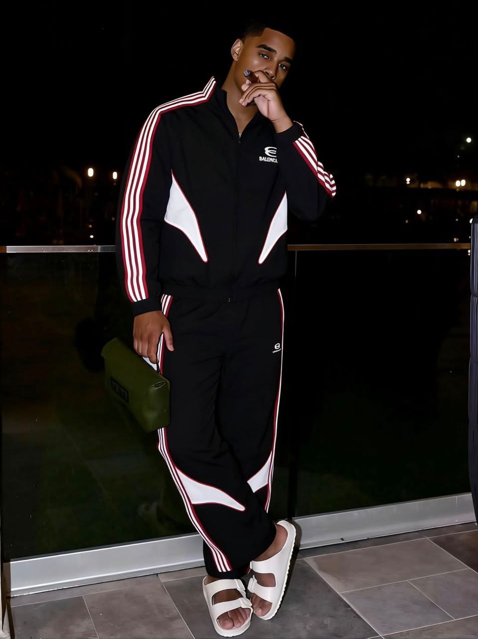 Young man posing in black Balenciaga tracksuit with red and white stripes on a balcony.