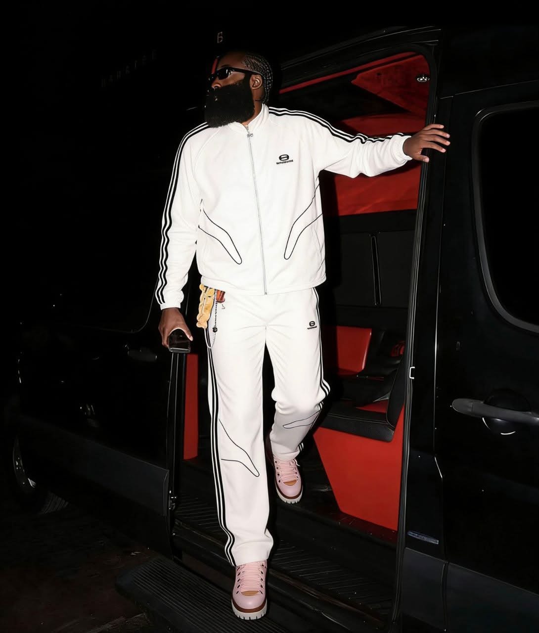 Man wearing white Balenciaga tracksuit with black stripes next to a vehicle at night.