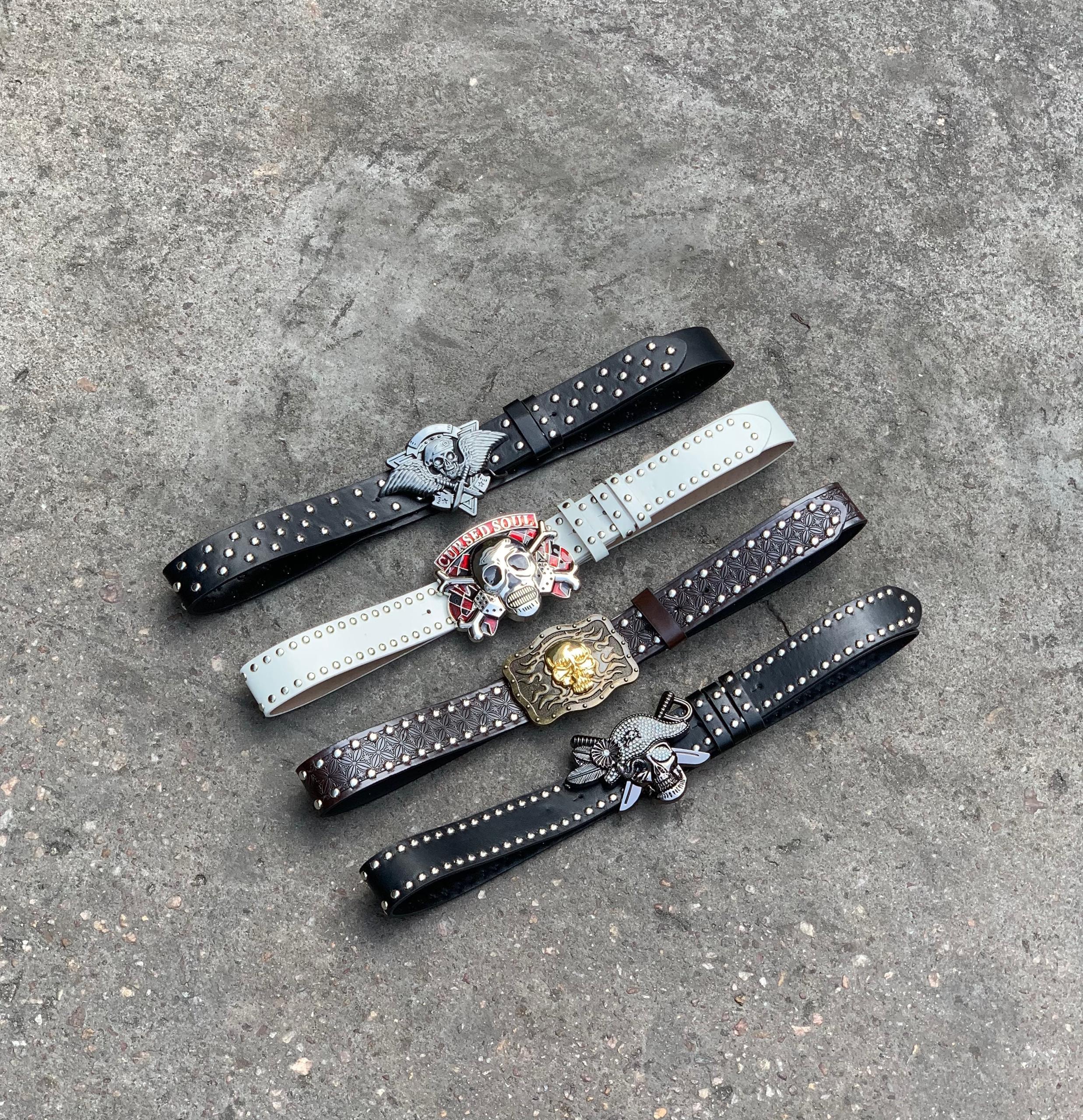 Four studded leather belts with skull and graphic buckles on gray surface.