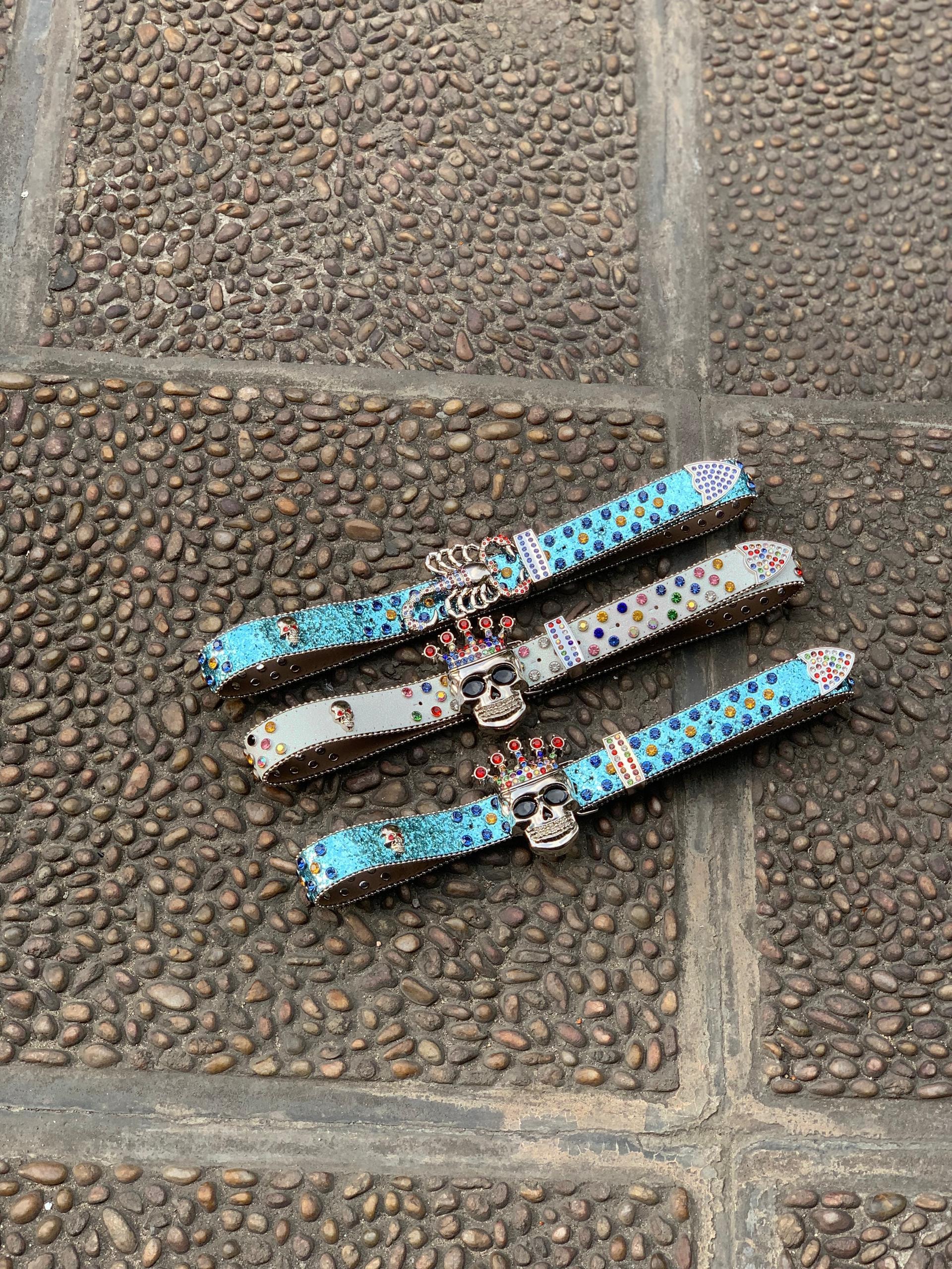 Assorted colorful rhinestone skull belts with crown buckles on stone pavement.