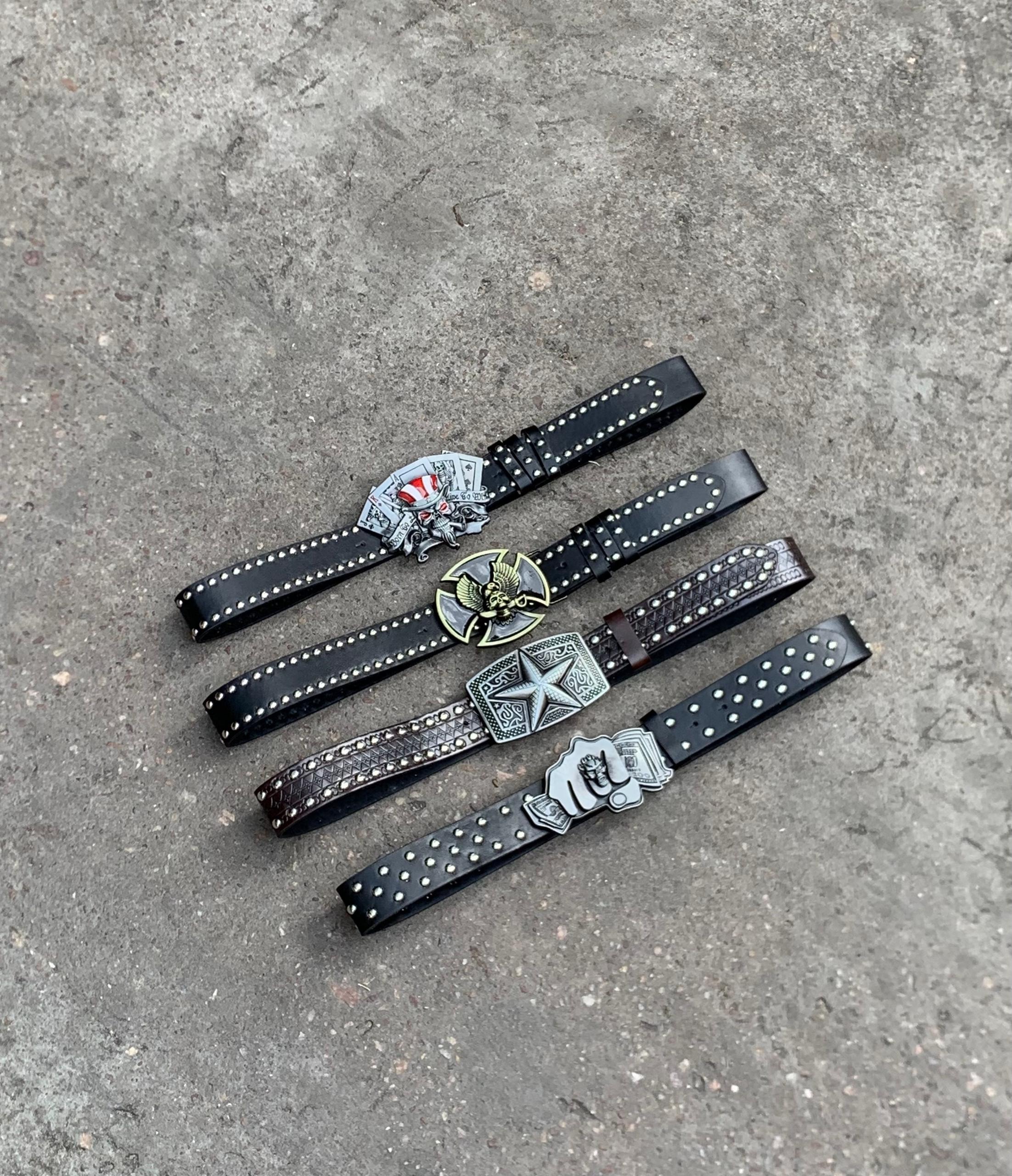 Four black leather belts with decorative buckles including skulls, cards, eagle and fist motifs on concrete.