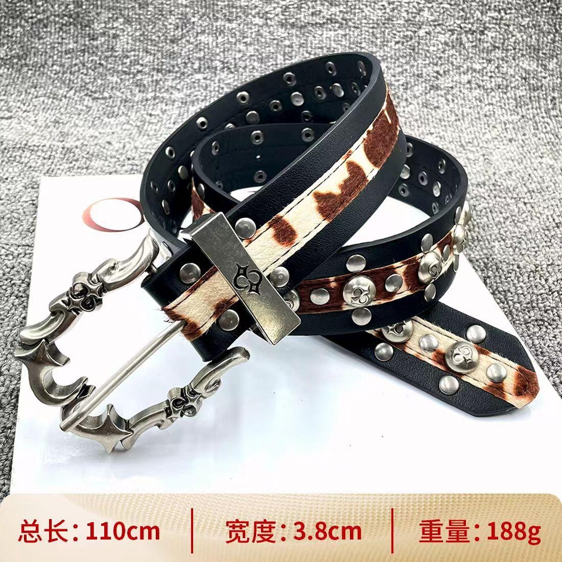 Black cow print leather belt with silver ornate buckle and studs on textured background.