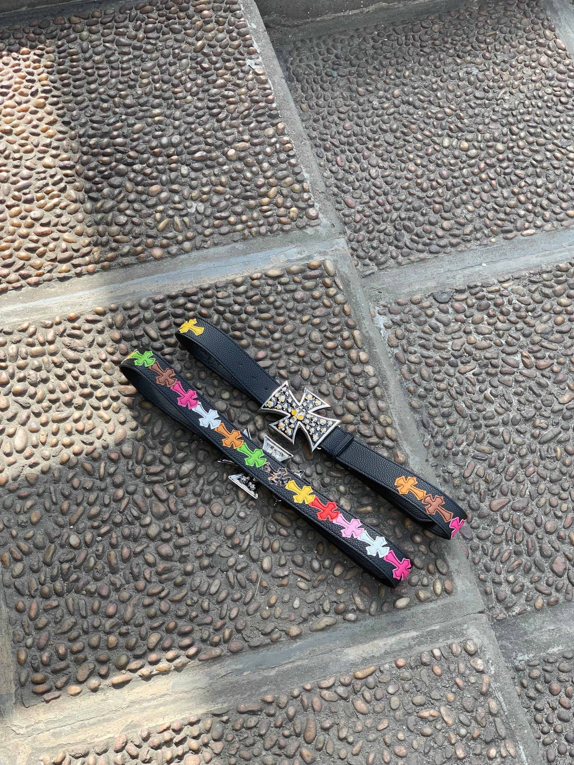 Black leather belts with colorful cross patterns and jeweled cross buckles on cobblestone.