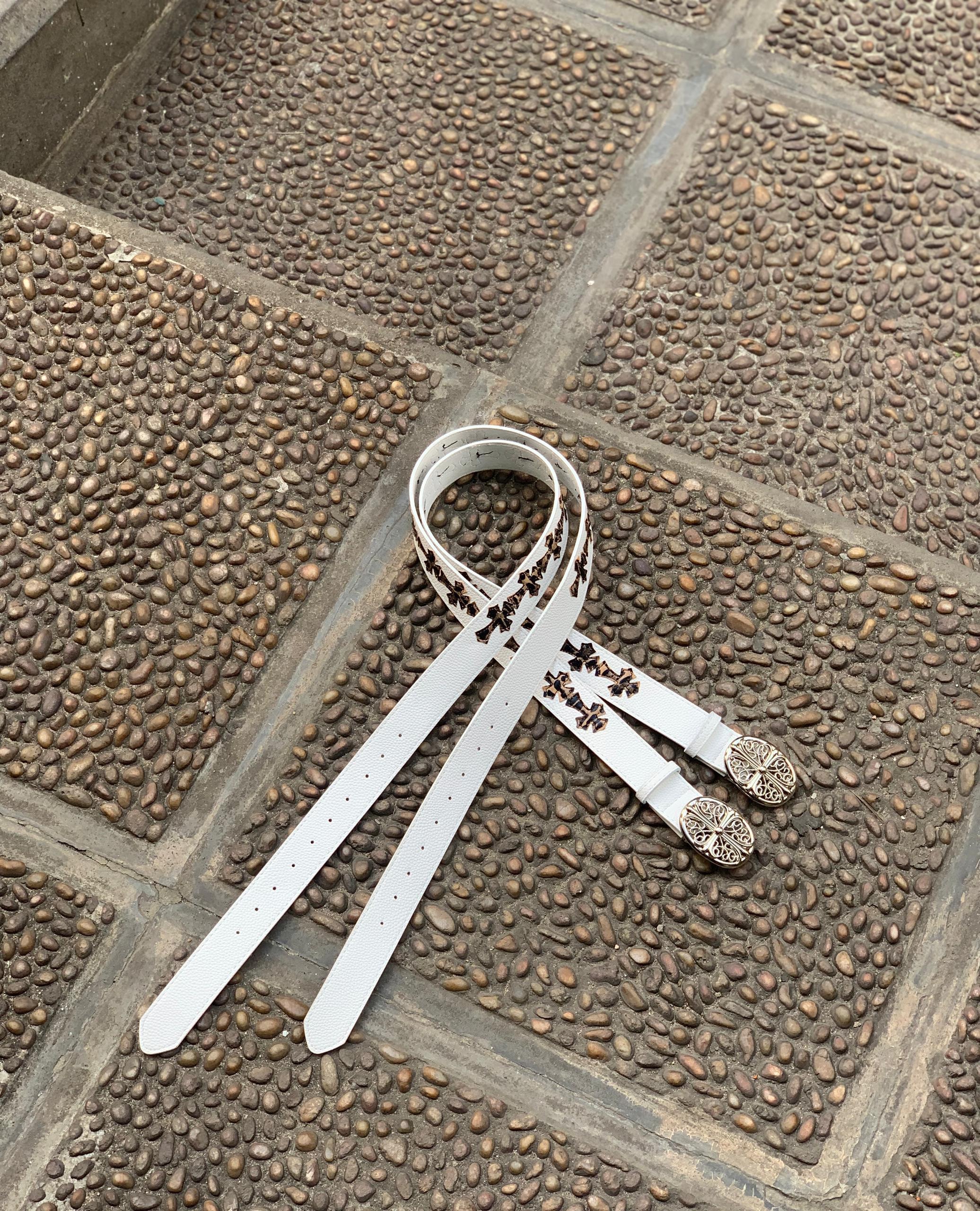 White leather belt with black cross patterns and ornate silver buckles on cobblestone.