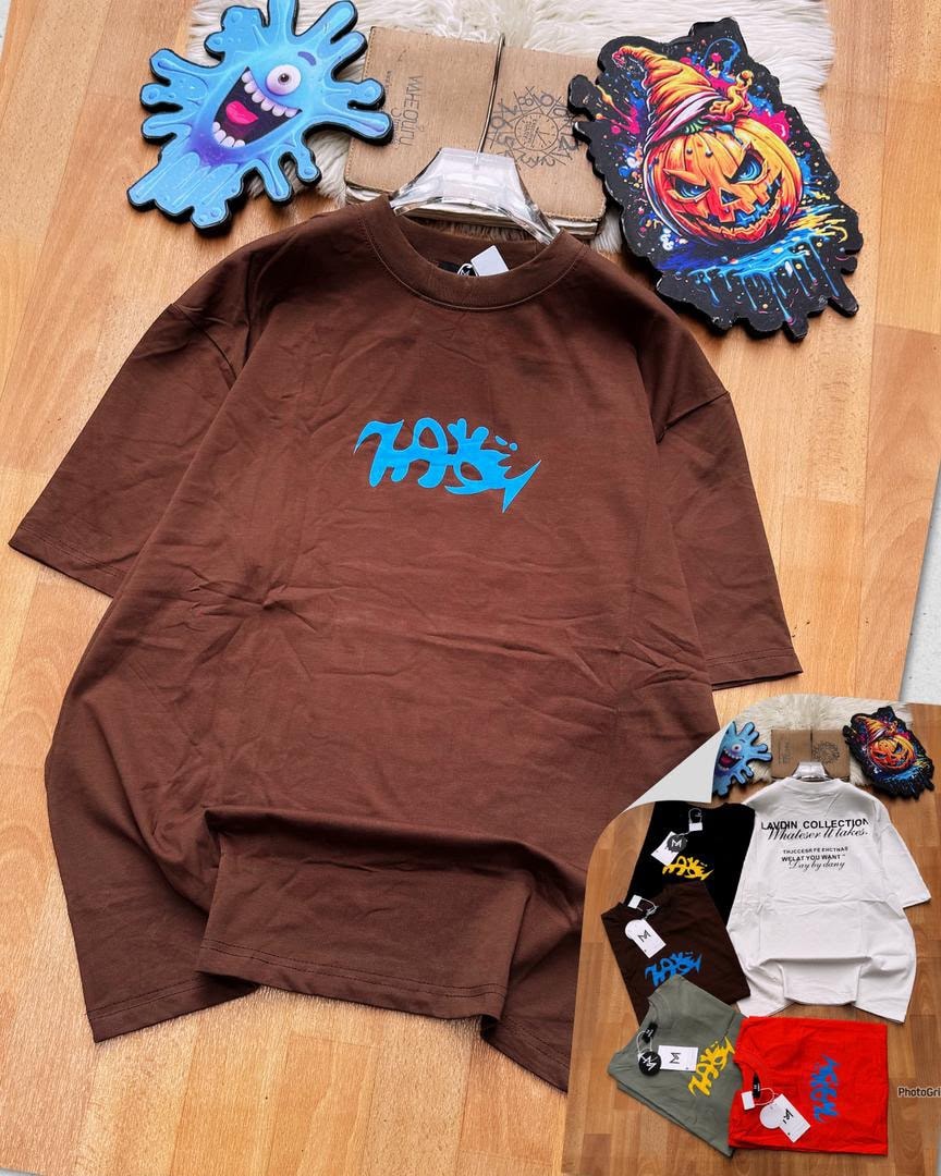 Brown oversized t-shirt with blue chest graphic, displayed on wooden floor with background items and color variants.